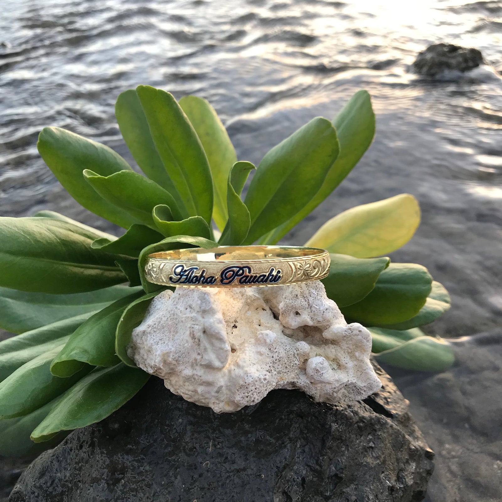 Aloha Pauahi 10mm yellow gold plated sterling silver Hawaiian heirloom bangle with black enamel script on coral among naupaka leaves by the water, handcrafted by Jewel Box Hawaii in Honolulu.
