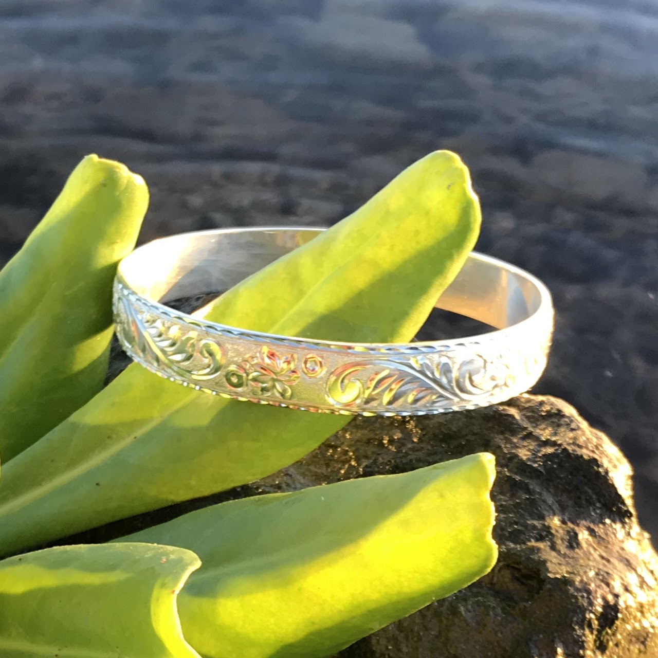 Aloha Pauahi 10mm solid gold Hawaiian heirloom bangle with hand engraved scrollwork on lava rock among green leaves, handcrafted by Jewel Box Hawaii in Honolulu.