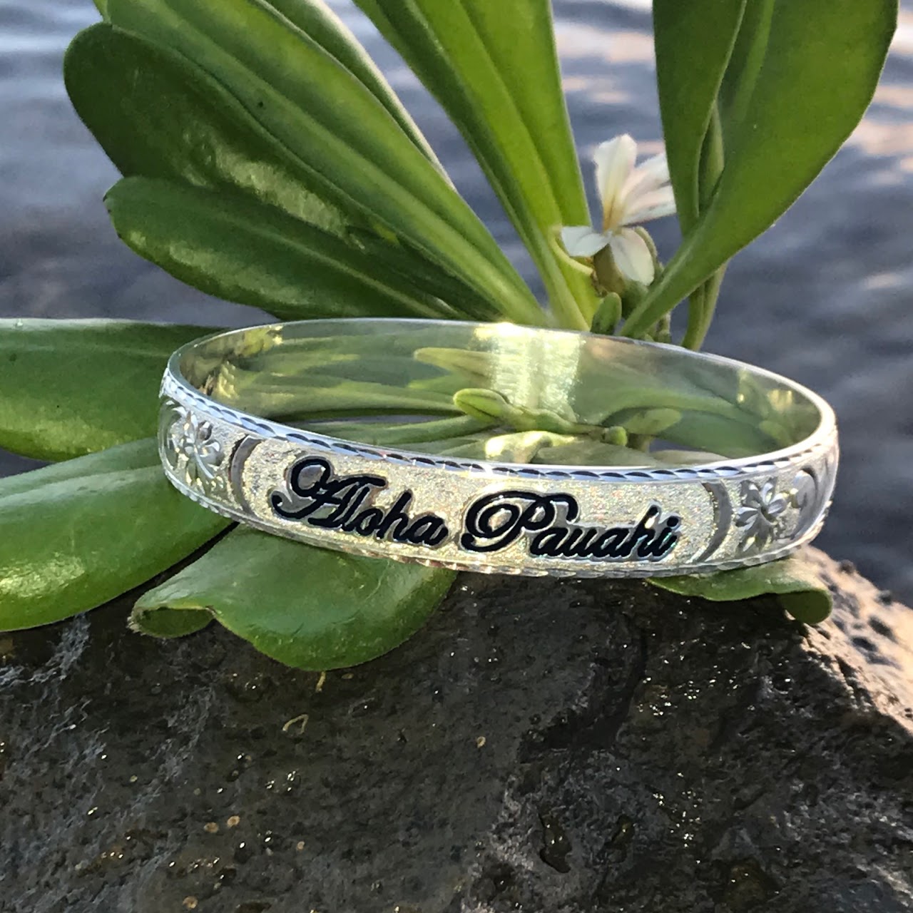 Aloha Pauahi 10mm sterling silver Hawaiian heirloom bangle with black enamel script on lava rock among naupaka leaves, handcrafted by Jewel Box Hawaii in Honolulu.