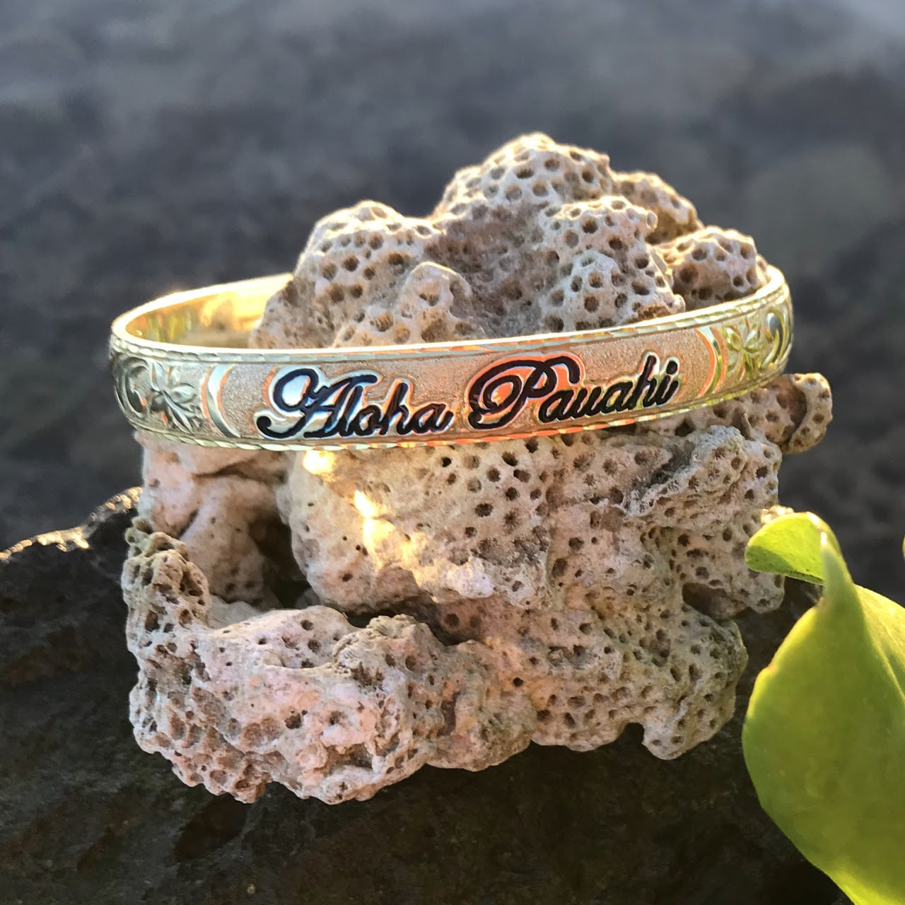 Aloha Pauahi 8mm yellow gold plated sterling silver Hawaiian heirloom bangle with black enamel script resting on coral, handcrafted by Jewel Box Hawaii in Honolulu.