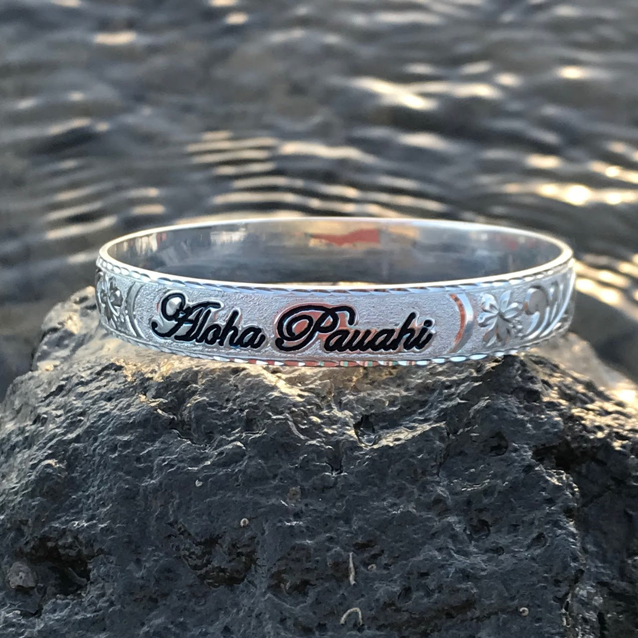 Aloha Pauahi 8mm sterling silver Hawaiian heirloom bangle with black enamel script on lava rock at the shoreline, handcrafted by Jewel Box Hawaii in Honolulu.