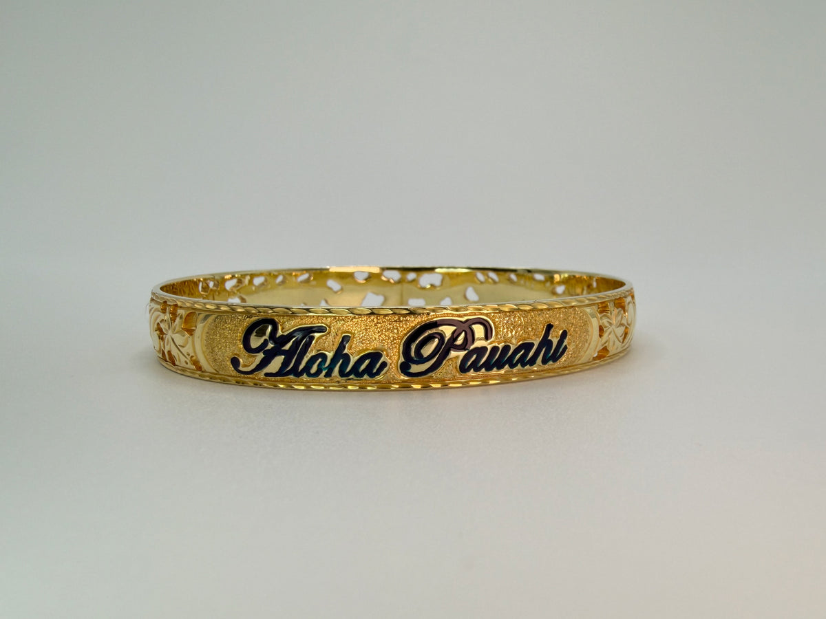 Aloha Pauahi gold Hawaiian heirloom bangle bracelet with wider pierced openwork band showing black-enamel 'Aloha Pauahi' nameplate front-facing center with plumeria cutouts flanking and sandblasted nameplate texture, handcrafted by Jewel Box Hawaii in Honolulu.
