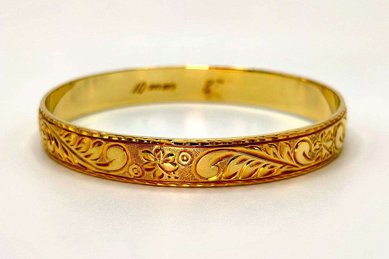Aloha Pauahi gold Hawaiian heirloom bangle with hand engraved scrolling leaf and flower motifs along the full band, seen from above on a plain light backdrop.