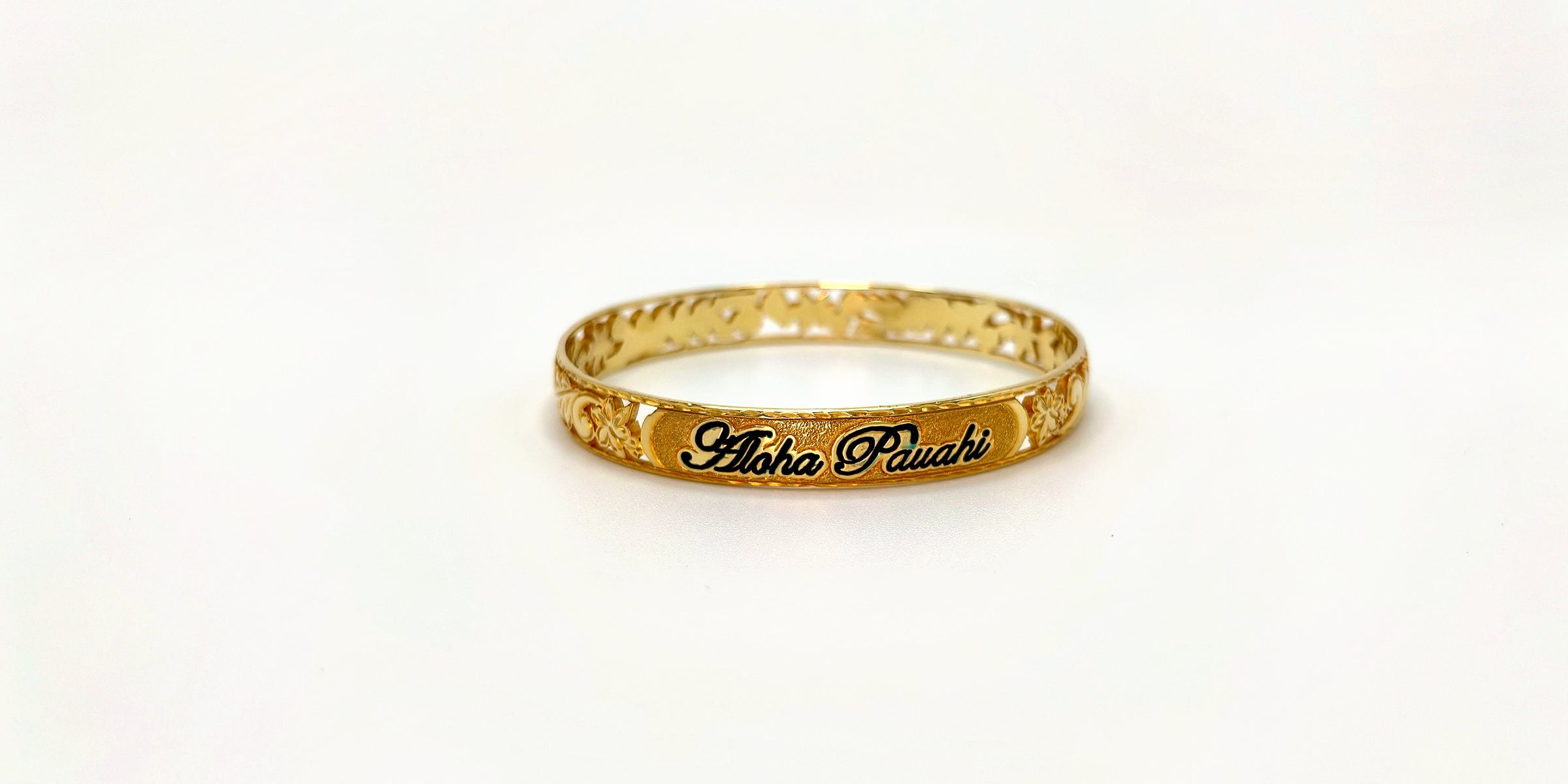 Aloha Pauahi gold Hawaiian heirloom bangle with the black enamel Aloha Pauahi nameplate centered on the front, on a plain light backdrop.