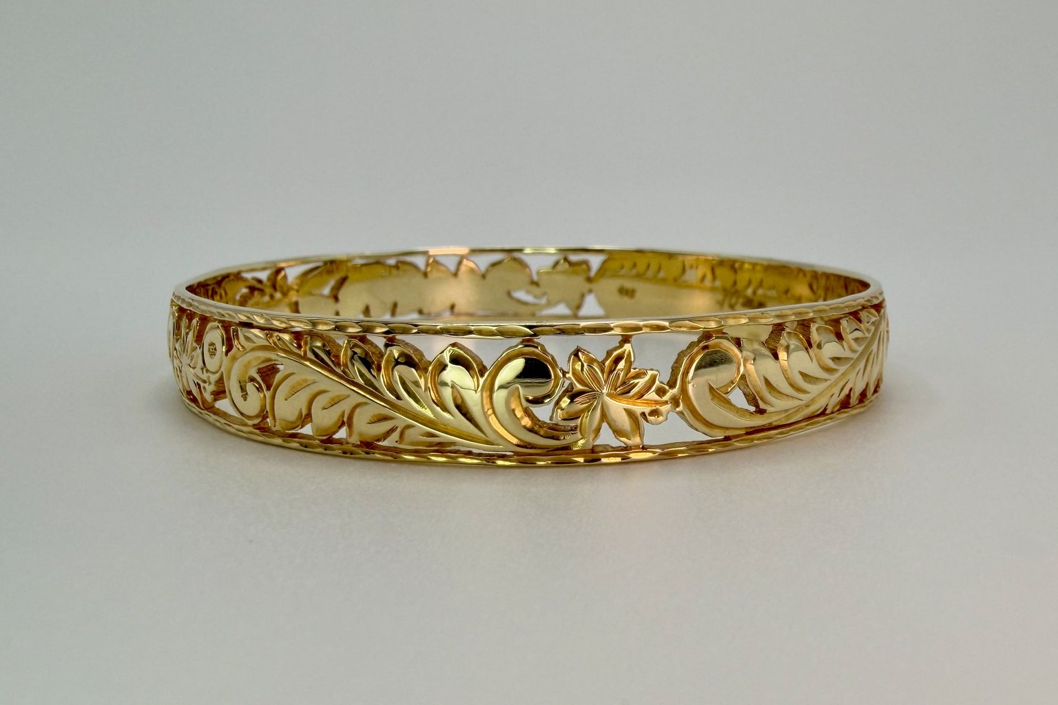 Aloha Pauahi gold Hawaiian heirloom bangle with open hand engraved leaf and blossom scrollwork, on a plain light backdrop.