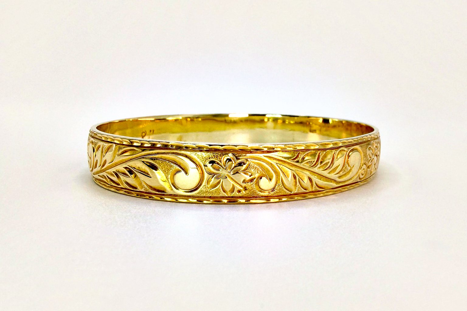 Aloha Pauahi gold Hawaiian heirloom bangle with a solid hand engraved band of leaves and a blossom, on a plain light backdrop.