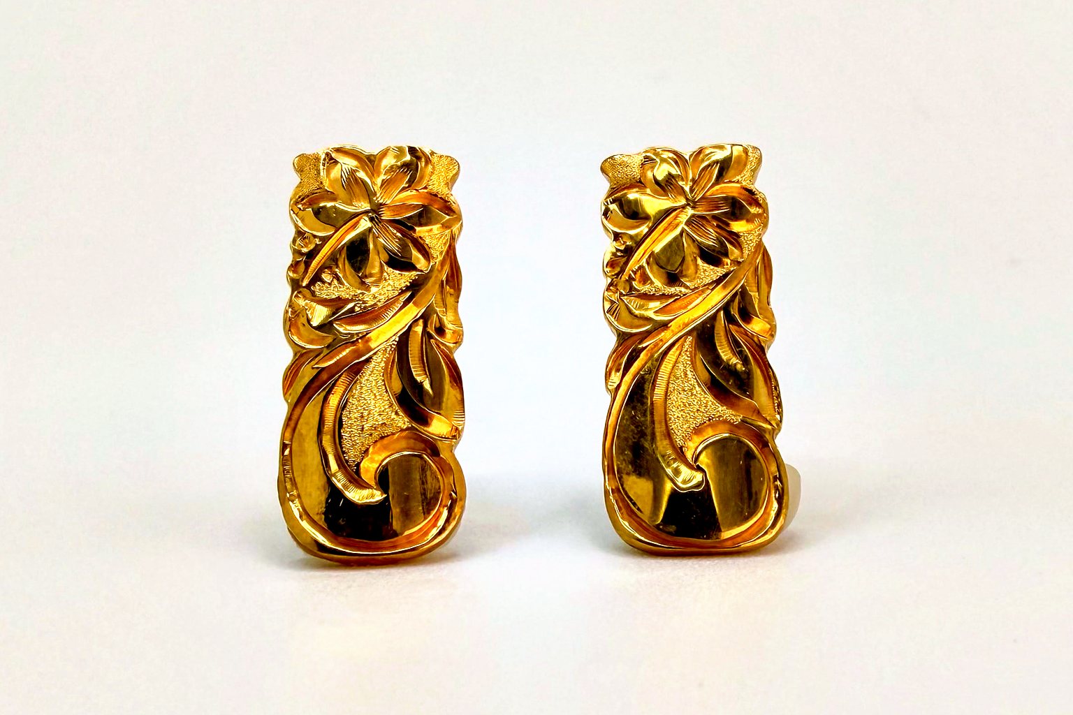 Pair of gold earrings engraved with a blossom and flowing scrolls over a textured ground, on a plain light backdrop.