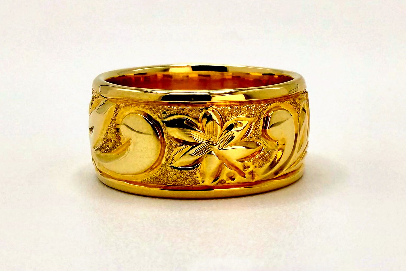 Aloha Pauahi gold Hawaiian heirloom ring with a raised flower and a scrollwork border, on a plain light backdrop.